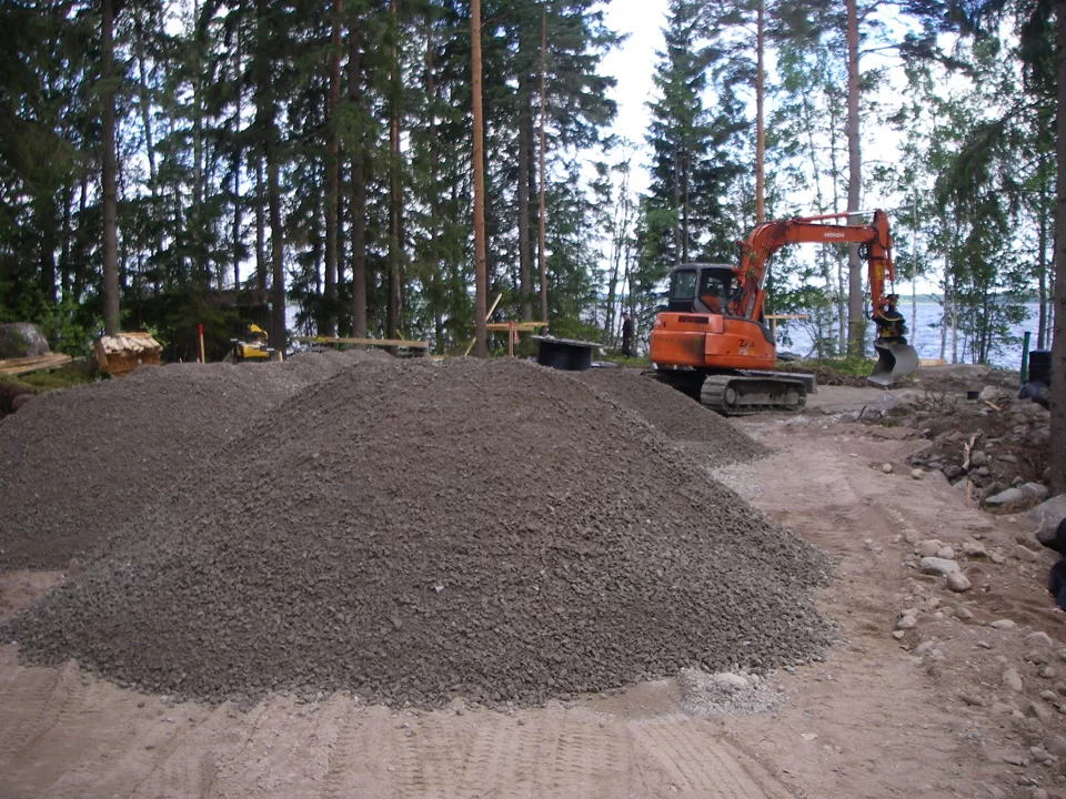 Hirsimökin maansiirtotyöt käynnissä rantatontilla – kaivinkone ja kaksi murskeläjää tulevan rakennuspaikan pohjalla.