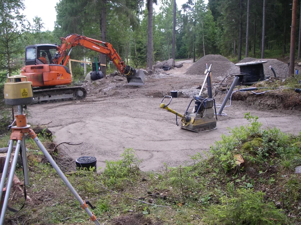 Hirsimökin perustusten pohjan tiivistys tärylevyllä – taustalla kaivinkone, sähkökaapelikela ja viemäriputkia, etualalla mittauslaite kolmijalalla.